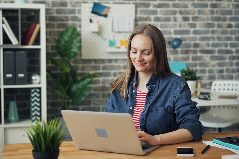 Een vrouw zit aan een bureau en werkt glimlachend op haar laptop.