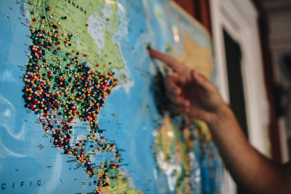 A world map with lots of pins in it and a person pointing at it.