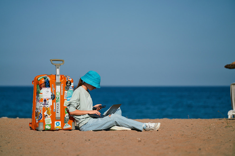 A woman sitting on the beach, leaning against her suitcase with a laptop in her lap and her phone in hand.