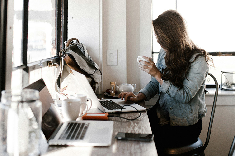 A woman sits at her laptop, typing, with a cup of coffee or tea in hand.