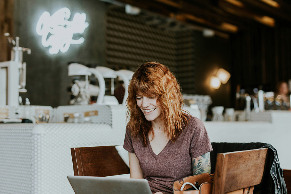 A woman smiles while looking at her laptop and sitting in a cafe.